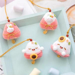 May include: Four handmade crocheted charms in the shape of a traditional Chinese sticky rice cake. Each charm features a different cute character, including a bunny, a sunflower, and two smiling faces. The charms are attached to gold chains and have a pink and white color scheme.