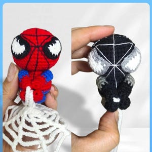 May include: Two small crocheted figures, one red and blue with a black web pattern and white eyes, the other black with a white web pattern and white eyes. Both figures are attached to a white crocheted web base and feature a round head design.