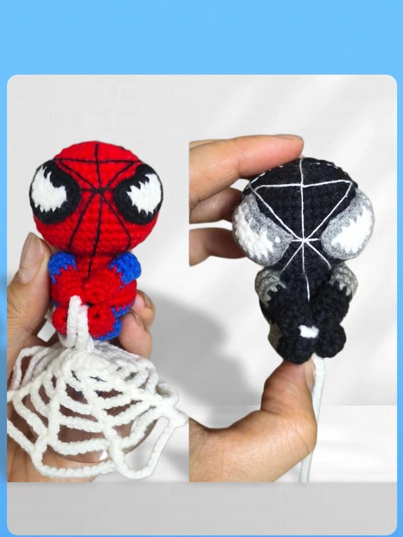 May include: Two small crocheted figures, one red and blue with a black web pattern and white eyes, the other black with a white web pattern and white eyes. Both figures are attached to a white crocheted web base and feature a round head design.