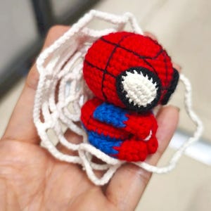 May include: A small, crocheted figure of a superhero with a red and blue body, a round red head with black web-like stitching, and large white eyes. The figure is surrounded by a white, circular, crocheted web-like structure made of yarn.