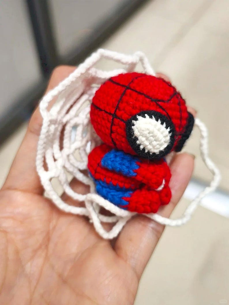 May include: A small, crocheted figure of a superhero with a red and blue body, a round red head with black web-like stitching, and large white eyes. The figure is surrounded by a white, circular, crocheted web-like structure made of yarn.