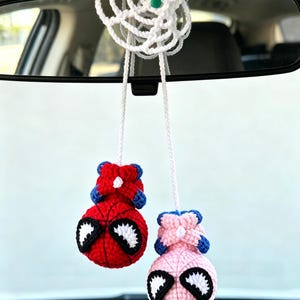 May include: A crochet car accessory featuring two spherical figures hanging from a white web-like structure. One figure is red and blue with black and white eyes, and the other is light pink and blue with black and white eyes, both suspended by white yarn.