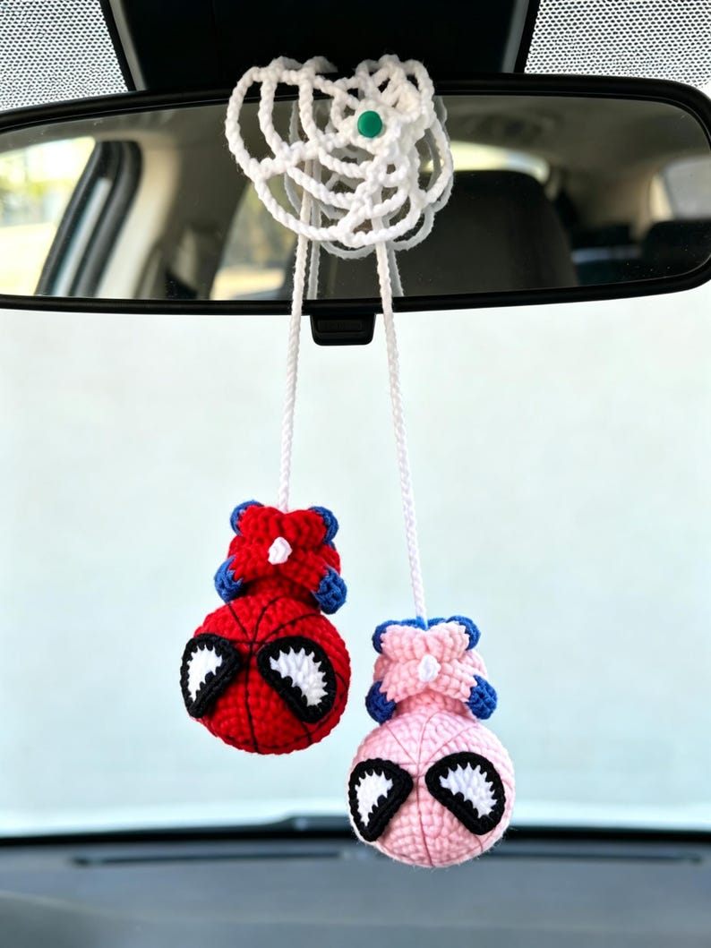 May include: A crochet car accessory featuring two spherical figures hanging from a white web-like structure. One figure is red and blue with black and white eyes, and the other is light pink and blue with black and white eyes, both suspended by white yarn.