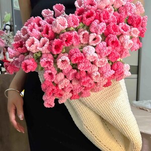 May include: A crocheted bag with a cream-colored base and a bouquet of pink and light pink crocheted flowers.