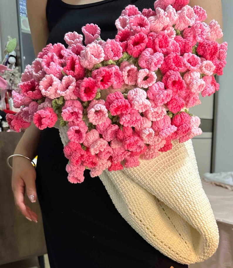May include: A crocheted bag with a cream-colored base and a bouquet of pink and light pink crocheted flowers.