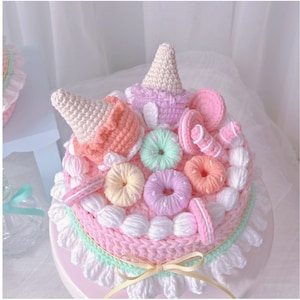 May include: A crocheted cake with pink, white and green frosting. The cake is decorated with crocheted doughnuts, ice cream cones, and other sweet treats.