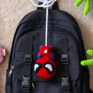 May include: A black backpack featuring a hanging crochet ornament of a red and blue character with large white eyes. The ornament is attached to a white crocheted web-like structure by a long white yarn string, centered on the backpack front.