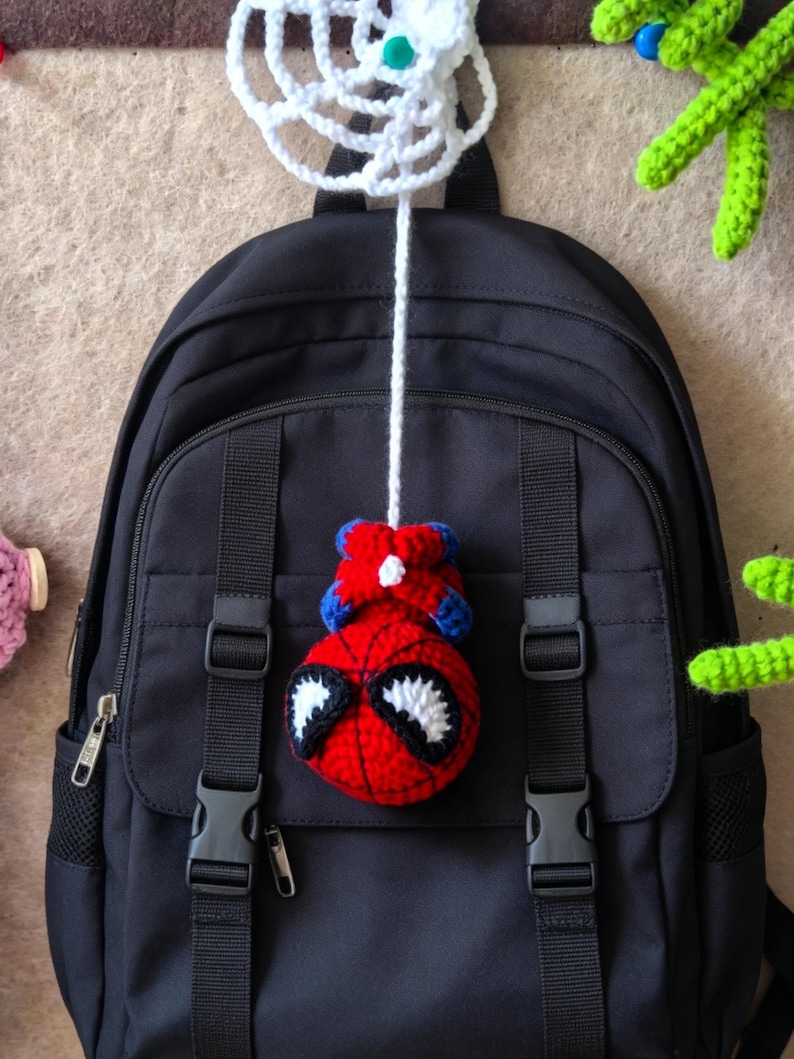 May include: A black backpack featuring a hanging crochet ornament of a red and blue character with large white eyes. The ornament is attached to a white crocheted web-like structure by a long white yarn string, centered on the backpack front.