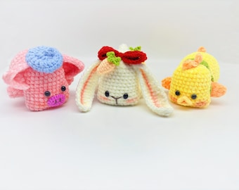 May include: Three crocheted stuffed animals: a pink pig wearing a blue hat, a white bunny with a red bow and a carrot, and a yellow chick with orange accents.