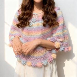 Puede incluir: Un poncho de crochet a rayas arcoíris pastel con detalles florales. El poncho presenta rayas horizontales rosas, amarillas, azules y lavanda. Flores de ganchillo se colocan alrededor de los bordes. La prenda es de tejido suave y abierto.