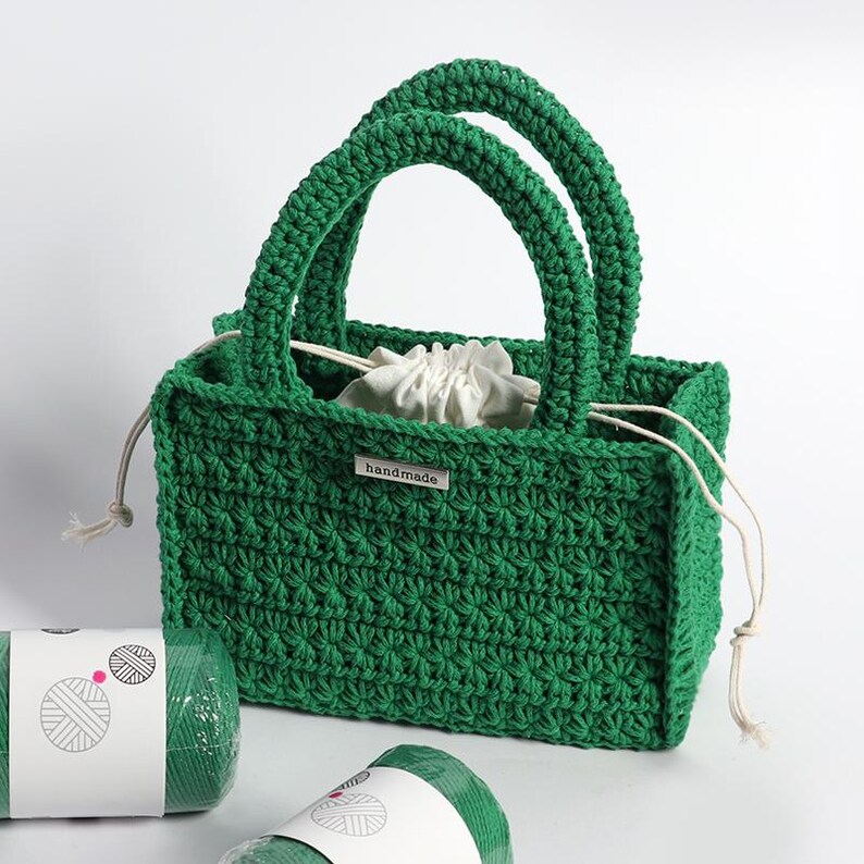 May include: A handmade green crochet tote bag with two handles and a white drawstring pouch inside. The bag has a textured stitch pattern and a small "handmade" tag. Two rolls of green yarn are in the foreground.