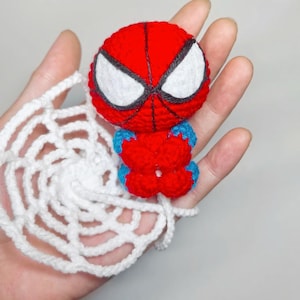 May include: A small, round, red and blue crocheted figure with large white eyes outlined in black, featuring a web-like pattern on the head. The figure is attached to a circular, white, crocheted spider web accessory made of yarn.