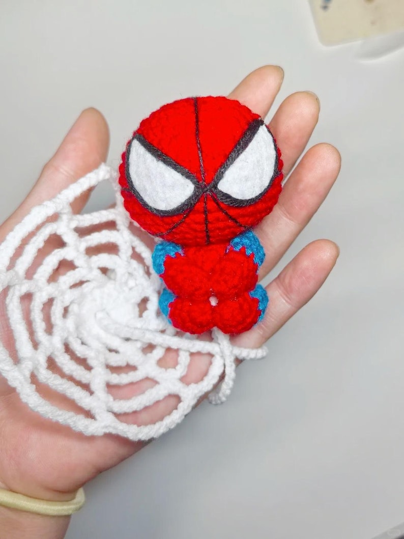 May include: A small, round, red and blue crocheted figure with large white eyes outlined in black, featuring a web-like pattern on the head. The figure is attached to a circular, white, crocheted spider web accessory made of yarn.