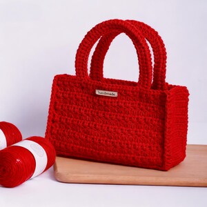May include: A handmade red crochet tote bag with two rounded handles. The bag has a rectangular shape and a small tag that reads "handmade". Two spools of red yarn are visible on the left side of the image.