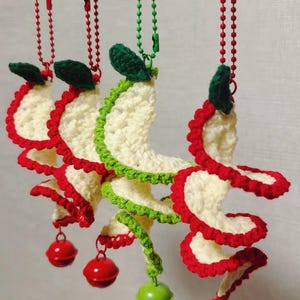 May include: Crocheted spiral ornaments in cream, red, and green, each with a small bell at the bottom. The ornaments are suspended from a metal bar by red and green beaded chains. The spiral design is reminiscent of a festive holiday decoration.