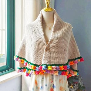 May include: A beige crochet shawl with a colorful flower trim. The shawl is on a dress form and has a tassel detail.
