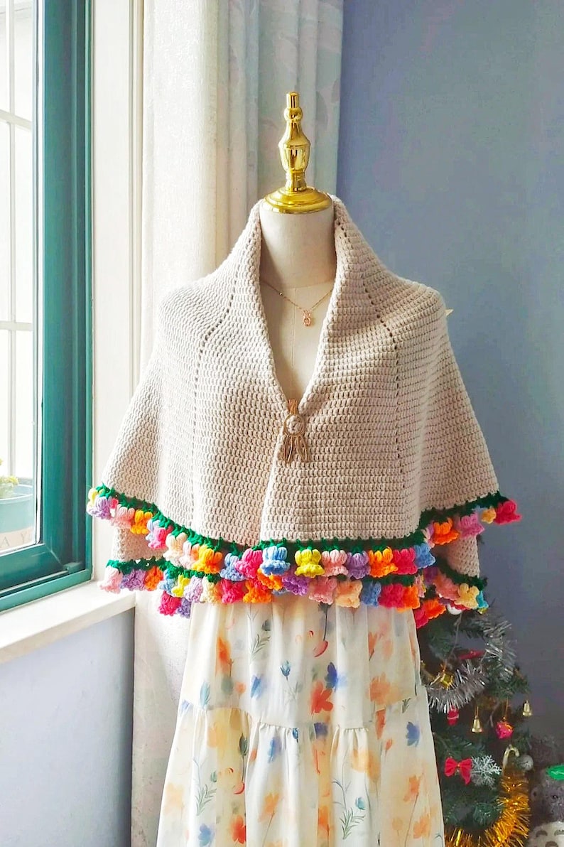 May include: A beige crochet shawl with a colorful flower trim. The shawl is on a dress form and has a tassel detail.