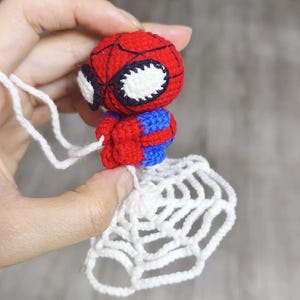 May include: A small, handmade crochet figure of a superhero character with a red and blue body, large white eyes with black outlines, and a black web pattern on the head. The figure is attached to a white crocheted spider web by a white yarn string.