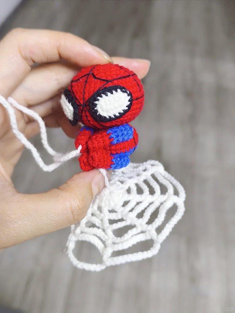 May include: A small, handmade crochet figure of a superhero character with a red and blue body, large white eyes with black outlines, and a black web pattern on the head. The figure is attached to a white crocheted spider web by a white yarn string.