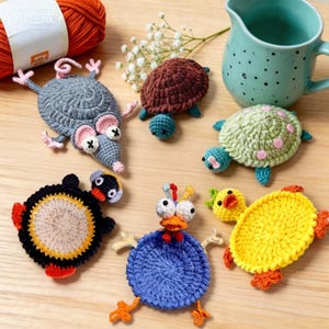 Pode incluir: Uma coleção de seis porta-copos de crochê em formato de animais, incluindo um rato cinza, uma tartaruga marrom, uma tartaruga verde-clara com manchas rosas, um pinguim preto e amarelo, um pássaro azul com bico vermelho e um pato amarelo, perto de uma caneca azul-petróleo.