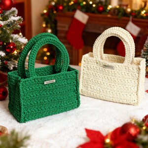 May include: Two handmade crochet tote bags. One is emerald green, the other is cream. Each has a rectangular shape, two handles, and a small metal tag that reads "handmade". The bags are on a white surface with Christmas decorations in the background.