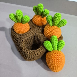 Op de afbeelding: Een handgemaakte gehaakte mand met wortelthema, met vier oranje wortels en groene toppen. De mand is bruin en heeft een getextureerd uiterlijk. De wortels zijn bovenop de mand geplaatst, wat een grillig en speels ontwerp creëert.