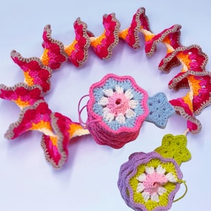May include: A collection of colorful, handmade crochet items. The pieces include a spiral garland of flower-like shapes in shades of pink, orange, and yellow, and several floral and star-shaped ornaments. The items are trimmed with a brown border.