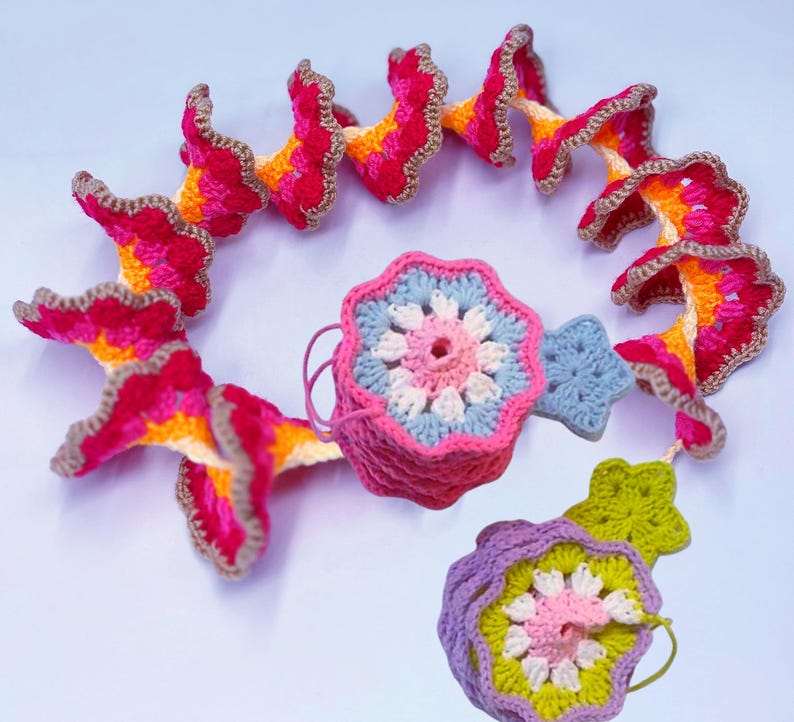 May include: A collection of colorful, handmade crochet items. The pieces include a spiral garland of flower-like shapes in shades of pink, orange, and yellow, and several floral and star-shaped ornaments. The items are trimmed with a brown border.
