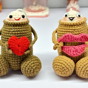 May include: Two handmade crochet figures with tan bodies, cream-colored heads, and round tan feet. One figure holds a red heart, the other holds pink lips. Both have black eyes and smiling faces. The figures are whimsical and decorative.