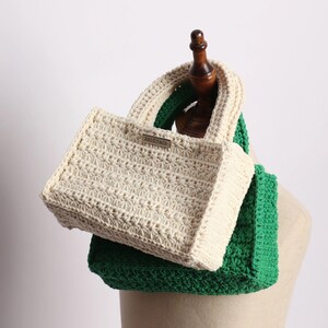 May include: A handmade, cream-colored crochet tote bag with a rectangular shape and a matching handle. The bag is placed on top of a green crochet scarf. A small metal tag on the bag reads "handmade".