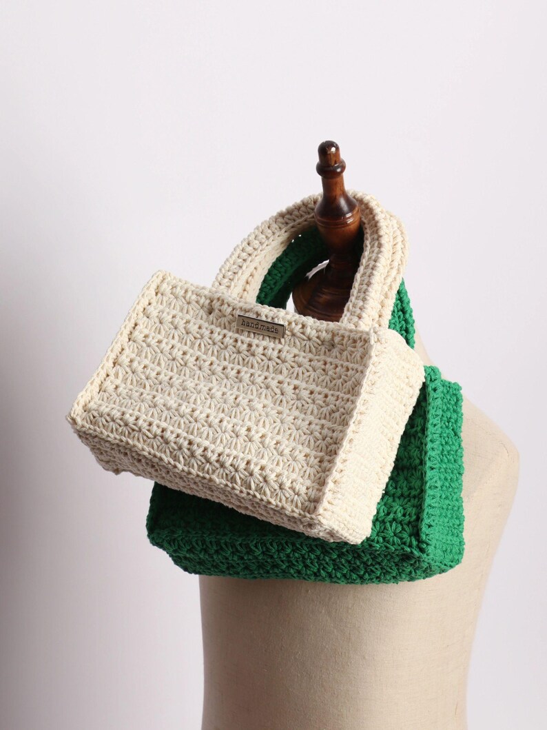 May include: A handmade, cream-colored crochet tote bag with a rectangular shape and a matching handle. The bag is placed on top of a green crochet scarf. A small metal tag on the bag reads "handmade".