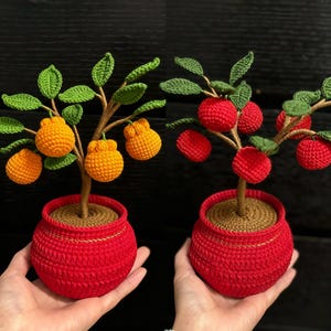 Könnte beinhalten: Zwei gehäkelte Bäume in roten Töpfen, einer mit orangen Früchten und der andere mit roten Früchten. Die Bäume haben braune Stiele, grüne Blätter und stehen vor einem dunklen Hintergrund. Die Töpfe haben einen goldenen Rand.