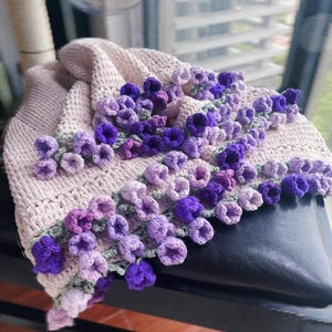 May include: A crocheted blanket with a pink background and a border of purple and lavender flowers. The blanket is folded and draped over a black chair.