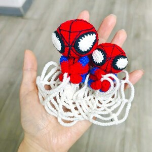 May include: Two small, handmade crochet figures featuring red and blue yarn with black web detailing and large white eyes. The figures are attached to a white crocheted web base, creating a cohesive set of two characters in a single piece.