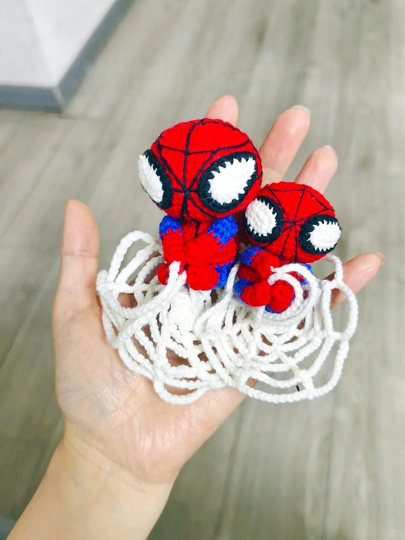 May include: Two small, handmade crochet figures featuring red and blue yarn with black web detailing and large white eyes. The figures are attached to a white crocheted web base, creating a cohesive set of two characters in a single piece.