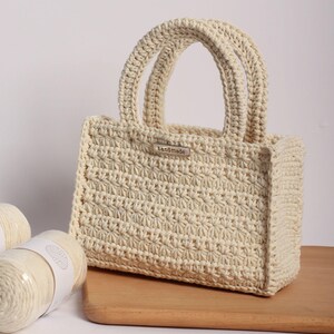 May include: Cream-colored, handmade crochet tote bag with two rounded handles. The bag features a textured stitch pattern and a small metal tag that reads "handmade". The bag is sitting on a wooden surface, with two spools of yarn visible.