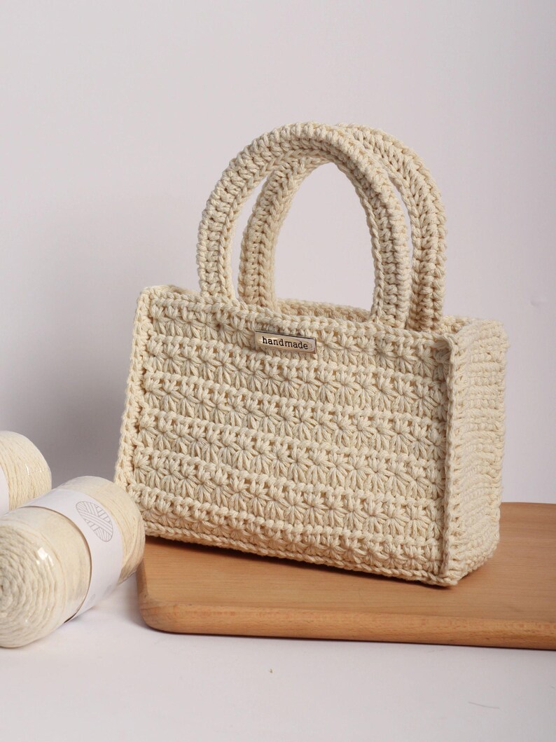 May include: Cream-colored, handmade crochet tote bag with two rounded handles. The bag features a textured stitch pattern and a small metal tag that reads "handmade". The bag is sitting on a wooden surface, with two spools of yarn visible.