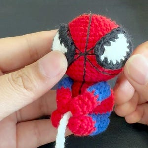 May include: A small, handmade crochet amigurumi figure of a superhero character with a red and blue yarn body. The figure features a round head with large white eyes outlined in black, a web-like pattern, and a long white yarn strand attached.