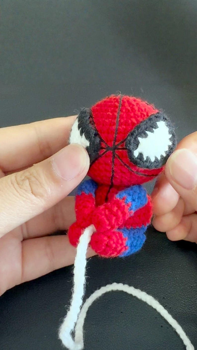 May include: A small, handmade crochet amigurumi figure of a superhero character with a red and blue yarn body. The figure features a round head with large white eyes outlined in black, a web-like pattern, and a long white yarn strand attached.