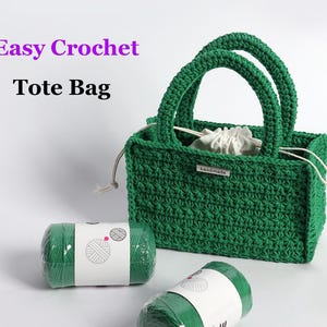 May include: A handmade green crochet tote bag with two handles, a drawstring closure, and a small "handmade" tag. Two rolls of green yarn are in the foreground. The text "Easy Crochet Tote Bag" is in the upper left corner.
