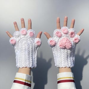 Peut inclure: Une paire de gants blancs au crochet en forme de pattes de chat. Les gants présentent des coussinets roses et sont présentés sur un fond neutre. Ces gants sont un accessoire unique.