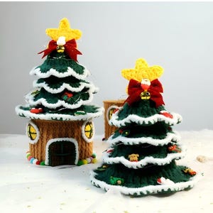 Puede incluir: Dos árboles de Navidad de crochet verde y blanco con estrellas amarillas y lazos rojos. El árbol más pequeño está sentado en una casa de crochet marrón.