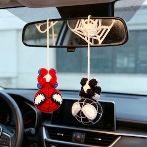 May include: Two crocheted yarn ornaments hanging from a car rearview mirror by white strings. One ornament is red and blue with large white eyes, and the other is black and white with large white eyes. A white crocheted spiderweb is attached above.