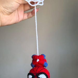 May include: A crocheted red and blue character head with large white eyes and black outlines, suspended by a long white yarn string from a circular white crocheted web-like piece. The character features a round red base with black web detailing.