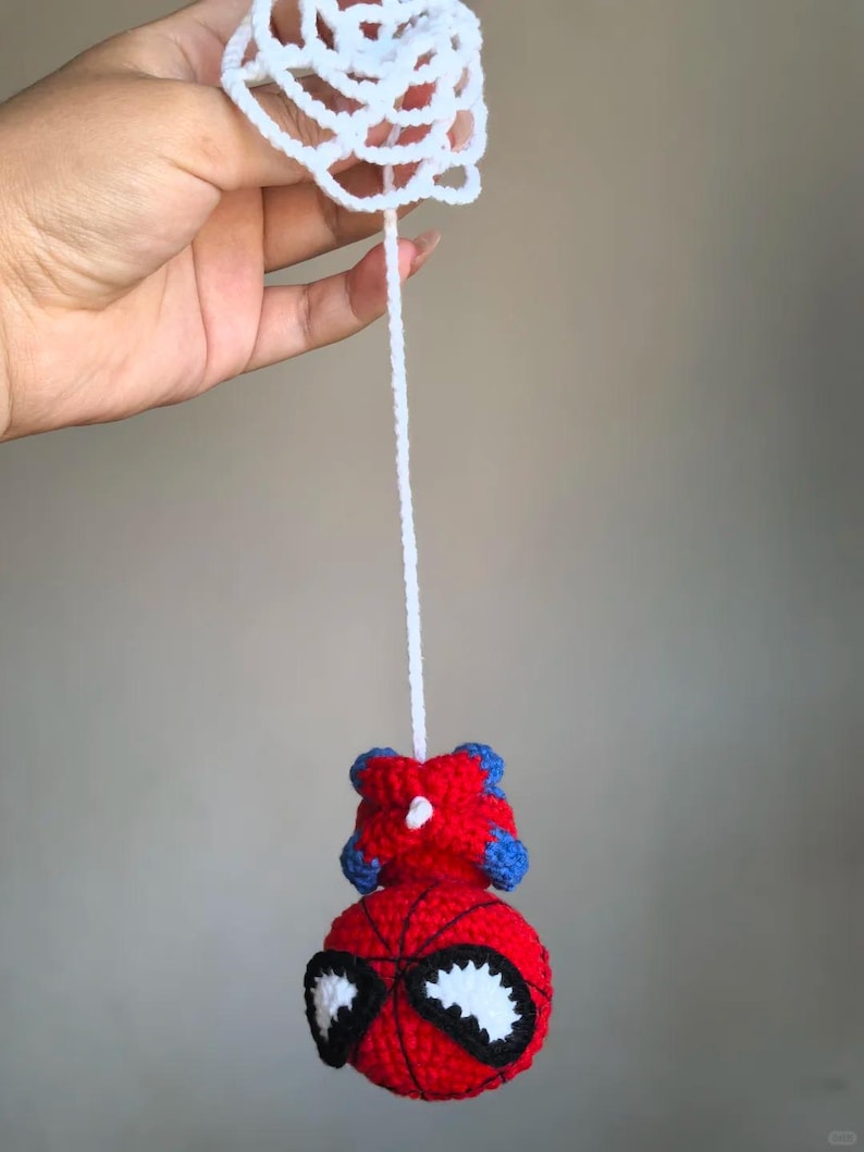 May include: A crocheted red and blue character head with large white eyes and black outlines, suspended by a long white yarn string from a circular white crocheted web-like piece. The character features a round red base with black web detailing.