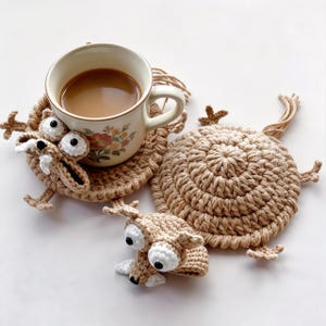 Peut inclure: Ensemble de dessous de verre crochetés beiges en forme de cerf. L'ensemble comprend un dessous de verre avec une tasse de café, un second dessous de verre et une petite tête de cerf. La tasse a un motif floral. Les dessous de verre ont de grands yeux blancs.