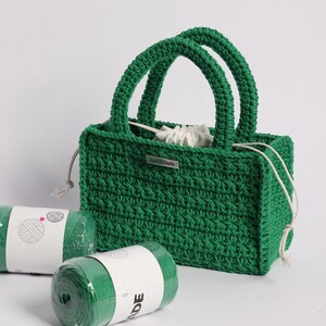 May include: A handmade green crochet tote bag with two rounded handles. The bag has a rectangular shape and a drawstring closure. Two rolls of green yarn are in the foreground. A small metal tag reads "handmade".