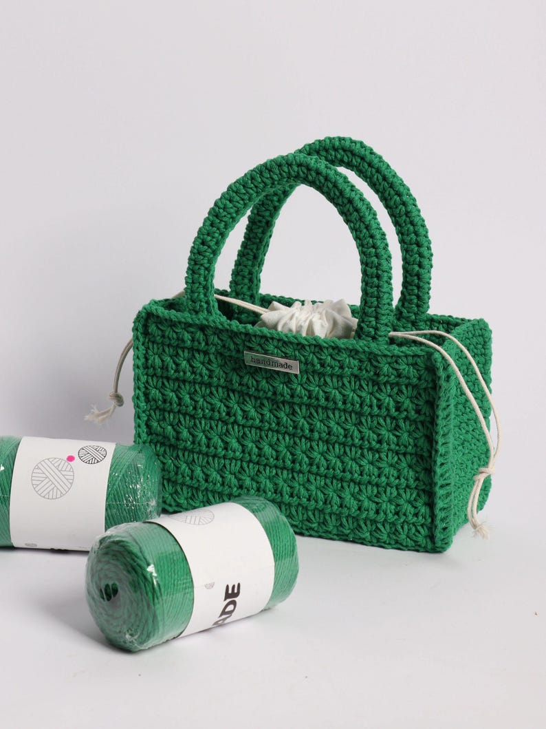 May include: A handmade green crochet tote bag with two rounded handles. The bag has a rectangular shape and a drawstring closure. Two rolls of green yarn are in the foreground. A small metal tag reads "handmade".