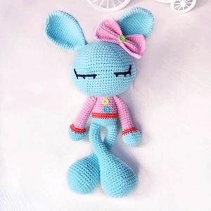 May include: A crocheted blue bunny with pink ears and a pink bow. The bunny has a pink and red sweater with colorful buttons. The bunny has closed eyes and a sweet expression.