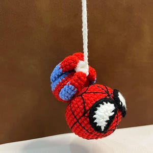 May include: A crocheted amigurumi figure of a superhero character with a round red head, black web-like stitching, and large white eyes. The figure is suspended by a white yarn string and features red and blue crocheted limbs attached to the top.